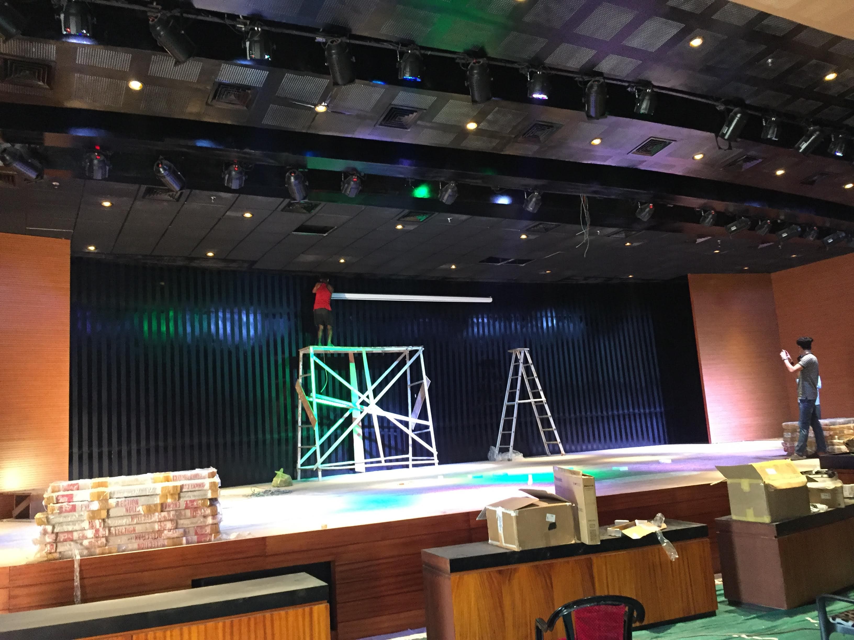 Multi-Purpose Hall Itanagar — stage with vertical timber-batten back-wall, screen install and overhead lighting rig in coloured wash