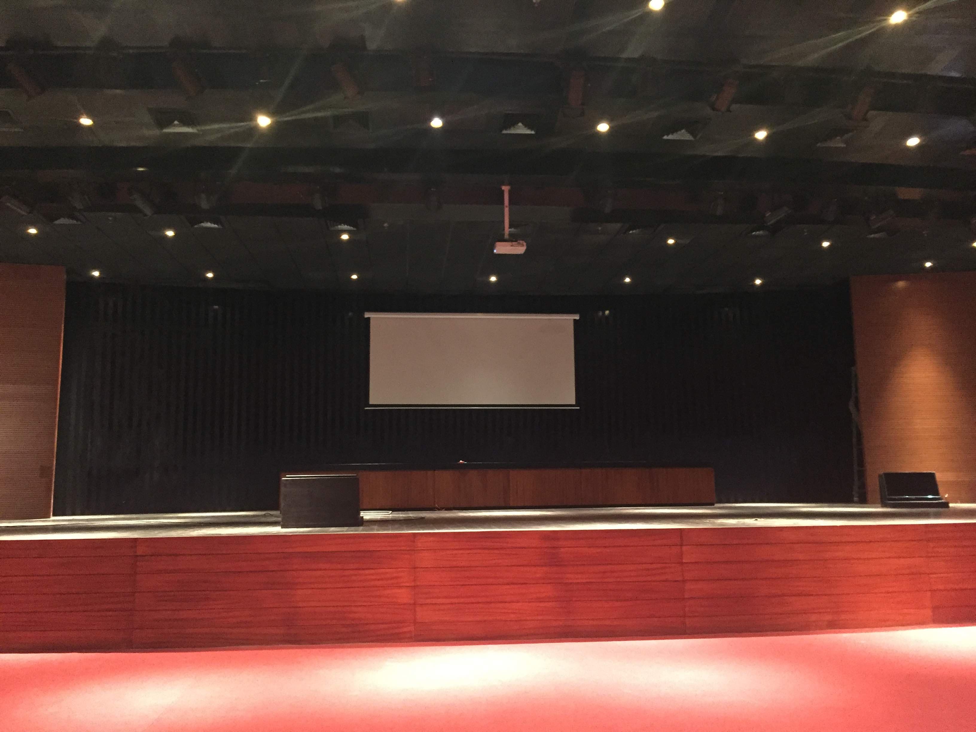 Multi-Purpose Hall Itanagar — stage with motorised projection screen down, floor loudspeakers and wooden podium under the lighting truss